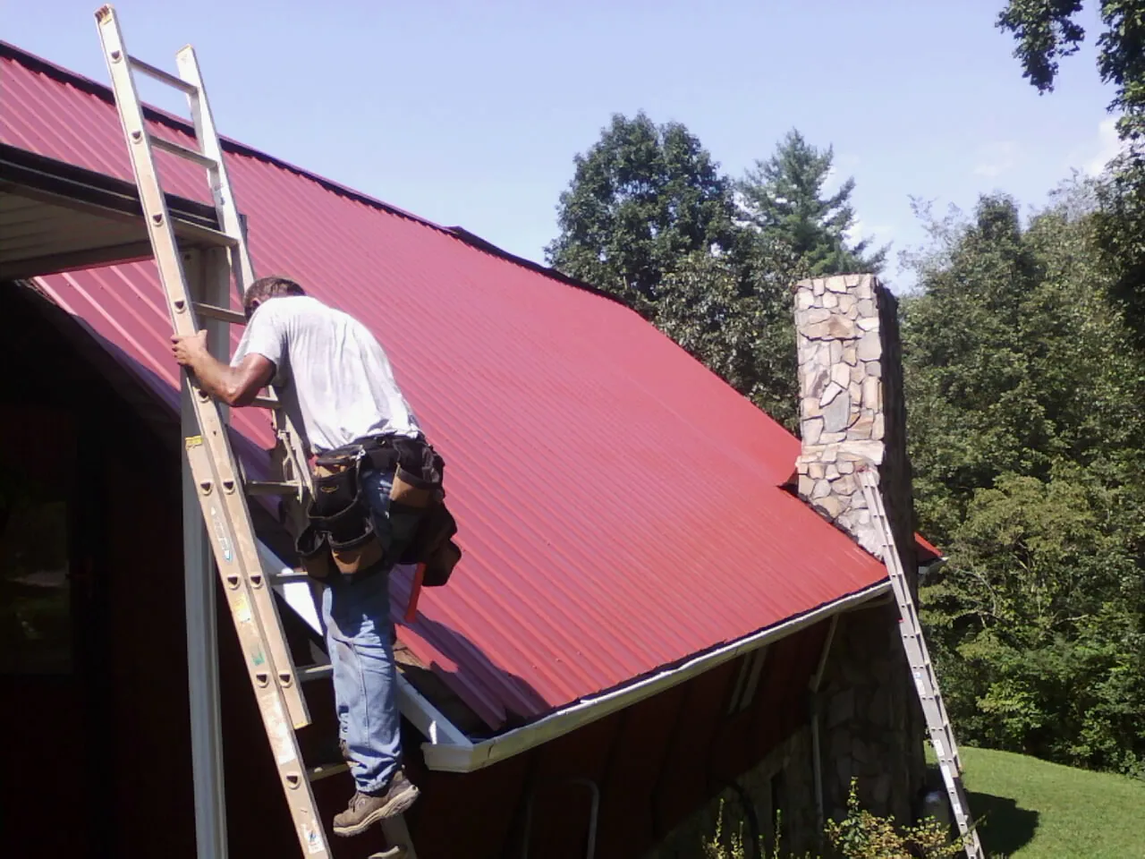 Roofer on a ladder beside a red metal roof and stone chimney — Soffit & Fascia Repair by Roofing Brothers in Cosmos